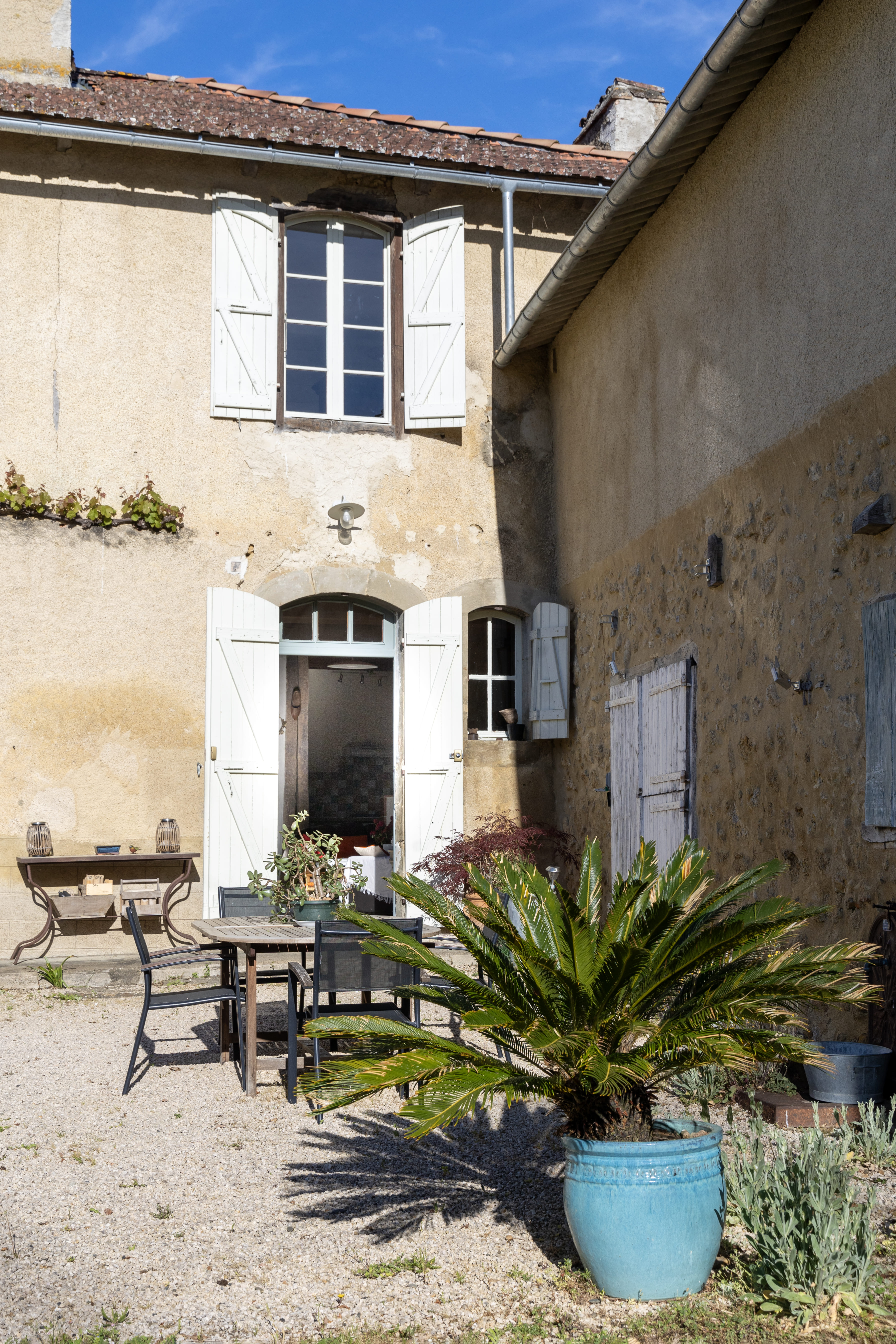 Vue extérieure d'un gîte en pierre avec des volets blancs, une table et des chaises en bois sur le devant, et une plante en pot dans le jardin.
